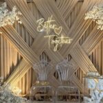 Décor événementiel élégant avec fond en tissu croisé beige, enseigne lumineuse Better Together, chaises transparentes sculptées et fleurs blanches en décoration