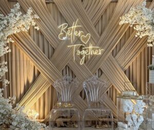Décor événementiel élégant avec fond en tissu croisé beige, enseigne lumineuse Better Together, chaises transparentes sculptées et fleurs blanches en décoration